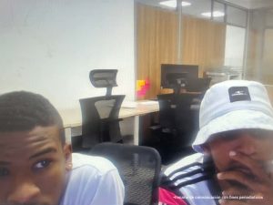 En la fotografía aparecen los rostros de dos hombres morenos, uno de ellos está vestido con camiseta blanca y el otro con camiseta blanca con rallas negras y un sombrero blanco. Ambas personas se encuentran sentadas en una oficina.