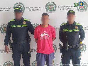 En la imagen aparece un hombre vestido con camiseta roja y pantalón corto azul, está de pie, con el rostro difuminado, los brazos esposados ​​a la espalda, acompañado de dos miembros uniformados de la Policía Nacional.