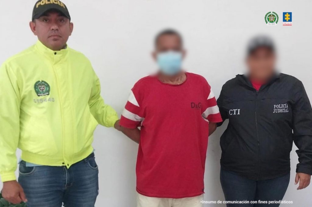 En la imagen está un hombre de pie, con el rostro difuso, los brazos atrás esposados, vestido con una camiseta roja y pantalón beige, custodiado por un funcionario del CTI y otro de la Policía Nacional.