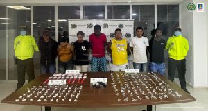 En la imagen se ve al grupo de capturados custodiados por dos uniformados de la Policía Nacional con un pendón de esa institución. Delante de ellos se observa una mesa de madera que tiene en la parte superior la droga y el arma incautada.