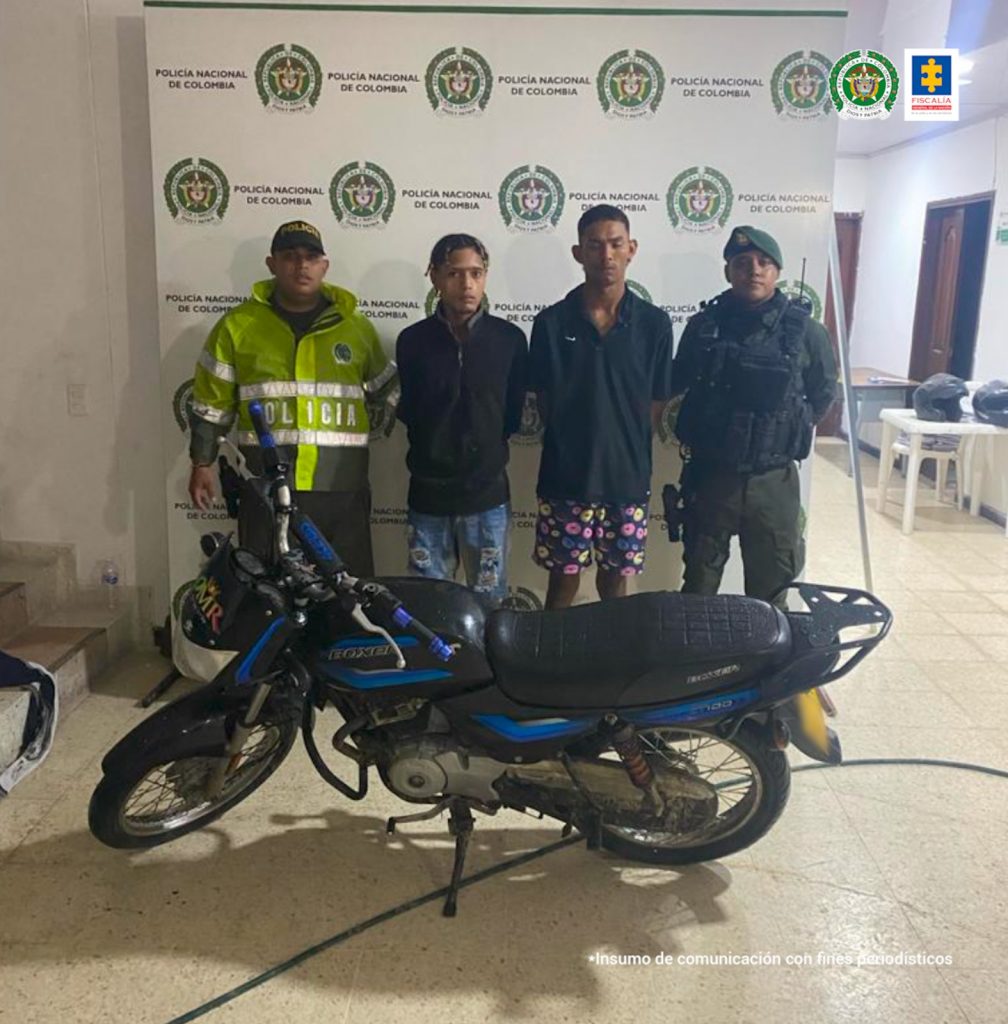En la fotografía se encuentra un hombre de pie, vestido de camiseta y bermuda blanca, con sandalias de rallas negras, blancas y rojas. Sus manos están esposadas y aparece acompañado de dos uniformados de la Policía Nacional.