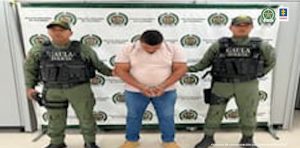 En la imagen está un hombre de camiseta rosada con jean azul y zapatos beige con los brazos hacia adelante esposado. Se encuentra custodiado por dos agentes de la Policía Nacional los cuales tienen un chaleco negro que dice Gaula.