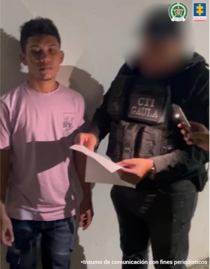 En la imagen, está un hombre joven, de pie, moreno, vestido con camiseta rosada y jean azul. Se encuentra con un uniformado del CTI que le lee los derechos de capturados.
