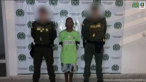 En la fotografía hay un hombre moreno, de estatura baja, se encuentra de pie con los brazos atrás, vestido de camiseta verde con bermuda de jean custodiado por dos agentes de la Policía Nacional.