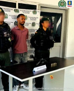 En la imagen está un hombre moreno de pie, con los brazos atrás, vestido de camiseta rosada, jean azul y tenis blanco, acompañado por dos agentes de la Policía Nacional. Delante de ellos hay una mesa con un arma de fuego incautada. 