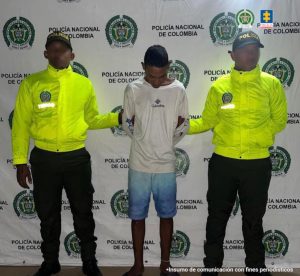 En la imagen está un un hombre delgado, de pie, con los brazos atrás, moreno, cabellos cortos negros, vestido con una camiseta blanca, una bermuda azul y sandalias negras. Se encuentra acompañado de dos uniformados de la Policía Nacional.