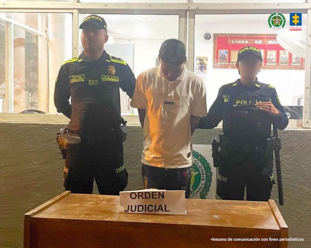 En la imagen está un hombre de pie, con los brazos atrás esposados, delgado, moreno con la cabeza agachada, tiene una gorra negra puesta y se encuentra vestido con una camiseta blanca y un jean negro con un roto en la pierna. Está acompañado de dos policías.