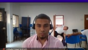 En la imagen está el medio cuerpo de un hombre joven, moreno, de cabellos cortos crespo, con barba, vestido con una camisa rosada. Se encuentra sentado con unos audífonos participando virtualmente de la audiencia en la que lo procesaron.