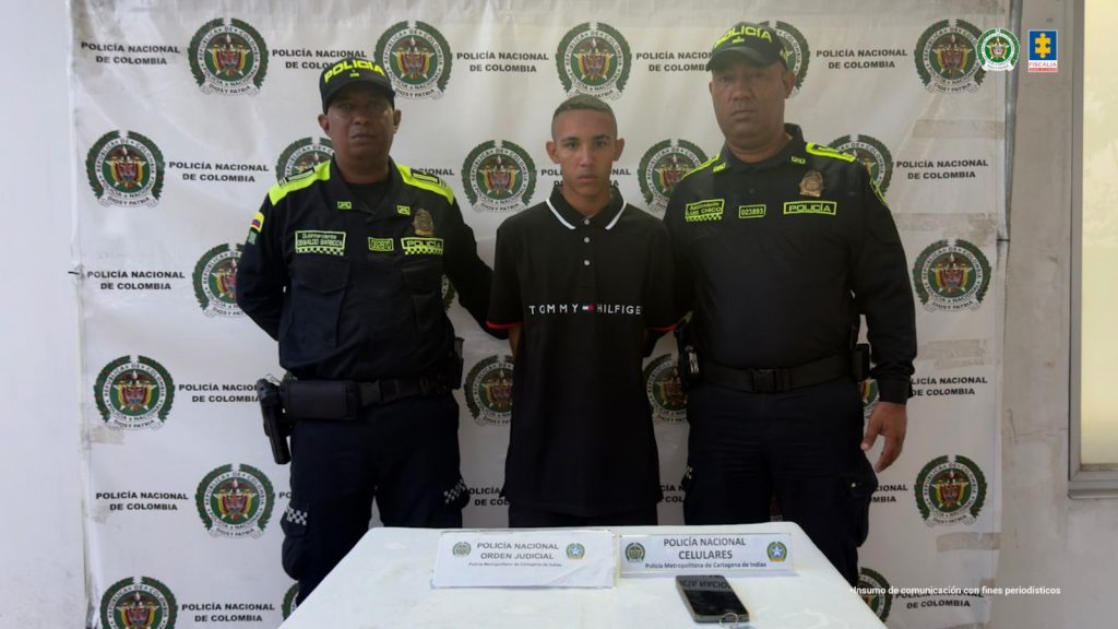 En la imagen está un hombre joven, de pie, de frente, trigueño, con los brazos atrás esposados, cabellos crespos cortos pintado de rubio, vestido con camiseta y pantalón negro y custodiado por dos policías. 