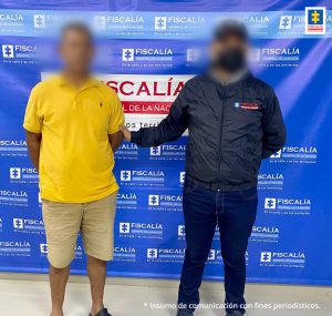 En la imagen hay un hombre de pie, con los brazos atrás, el rostro difuso, vestido de bermuda beige, camiseta amarilla y zapatos negros, custodiado por un funcionario del CTI. Detrás de ellos hay un pendón azul de la Fiscalía General de la Nación.