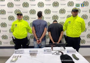En la imagen están dos hombres de pie, de espalda, con los brazos esposados, uno vestido con camiseta y jean azul y el otro con camiseta negra. Se encuentran custodiado por dos uniformados de la Policía Nacional. Delante de ellos hay una mesa con el fusil, el arma y las municiones incautadas.