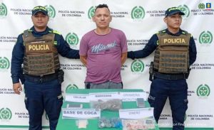 En la foto está un hombre de pie, de frente, trigueño, con los brazos atrás esposados, cabellos negros, contextura mediana, camiseta morada y sudadera gris. A su lado se encuentran dos policías custodiándolo.