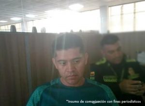 En la imagen está el rostro de un hombre delgado, trigueño, cabellos negros, vestido con una camiseta verde y se encuentra al interior de una oficina asistiendo virtualmente a la audiencia en la que se le procesó.