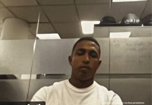 En la imagen está el rostro de un hombre moreno, joven, cabellos negros crespos cortos, está vestido con una camiseta blanca y se encuentra al interior de una oficina asistiendo virtualmente a la audiencia en la que se le procesó.