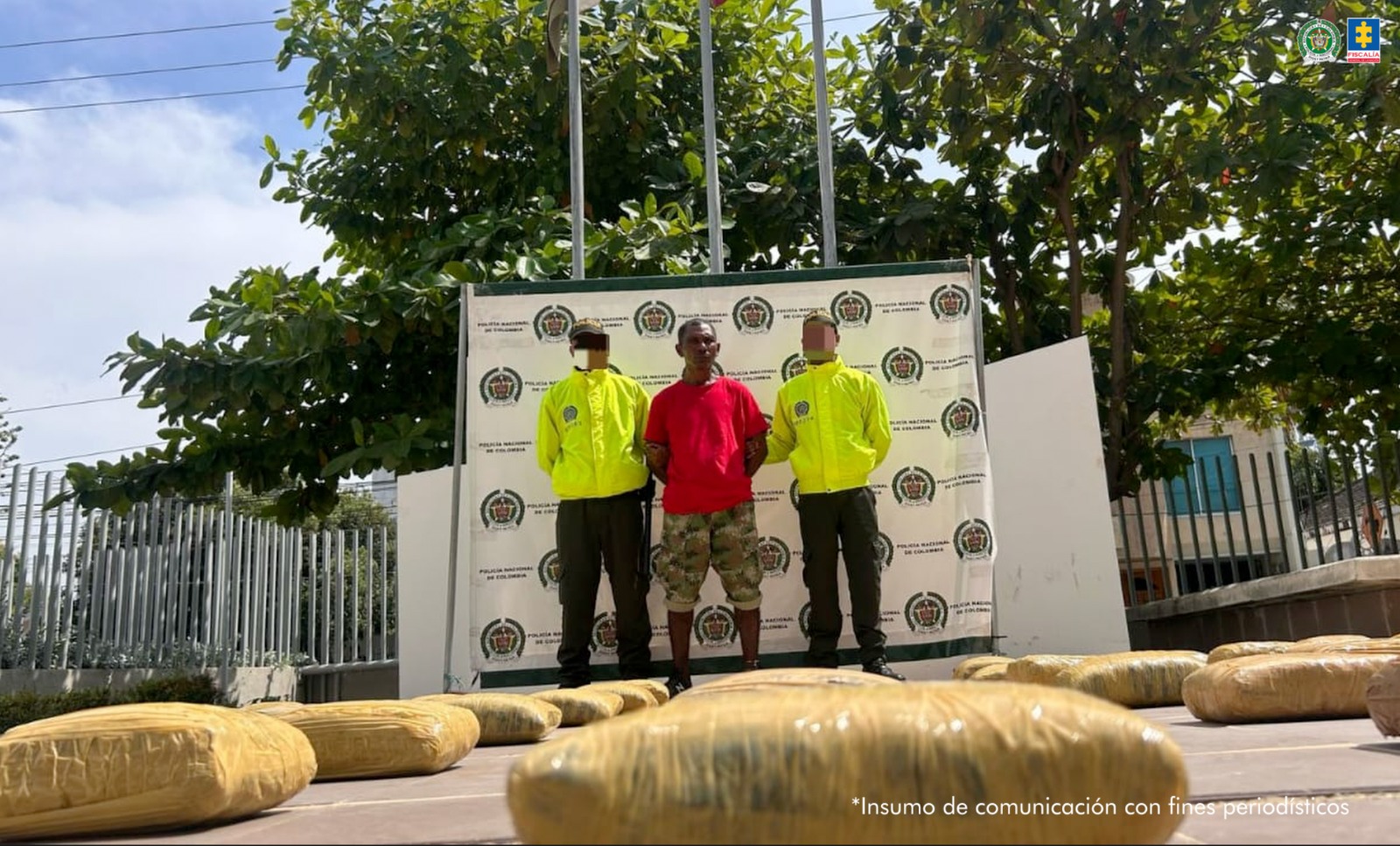 En la imagen aparece un hombre moreno de pie, con los brazos atrás esposados, vestido con una camiseta roja y un pantalón camuflado verde, estilo militar. Se encuentra acompañado por dos policías. Delante de él se ve la droga que se le incautó.