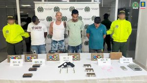 En la imagen aparecen cuatro hombres de pie con los brazos atrás, acompañados por dos agentes de la Policía Nacional. Delante de ellos está una mesa con la droga y armas incautadas. Detrás de ellos hay un pendón de la Policía Nacional.