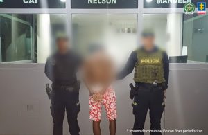 En la imagen está un hombre de espalda con los brazos esposados, moreno, viste una bermuda roja con flores blancas y unos tenis blancos. Se encuentra con dos uniformados de la Policía Nacional en un CAI