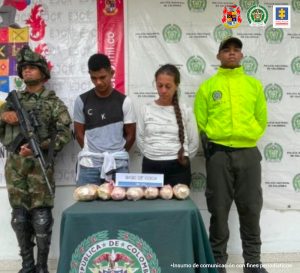 En la fotografía están un hombre de pie con la cabeza agachada y brazos atrás vestido de camiseta gris con jean azul. A su lado esta una mujer de cabellos largos con una trenza vestida de camiseta blanca y pantalón negro. Ellos se encuentran acompañados por un uniformado del Ejército Nacional y otro de la Policía Nacional. Delante de ellos hay una mesa con mantel verde donde se encuentra la droga incautada.