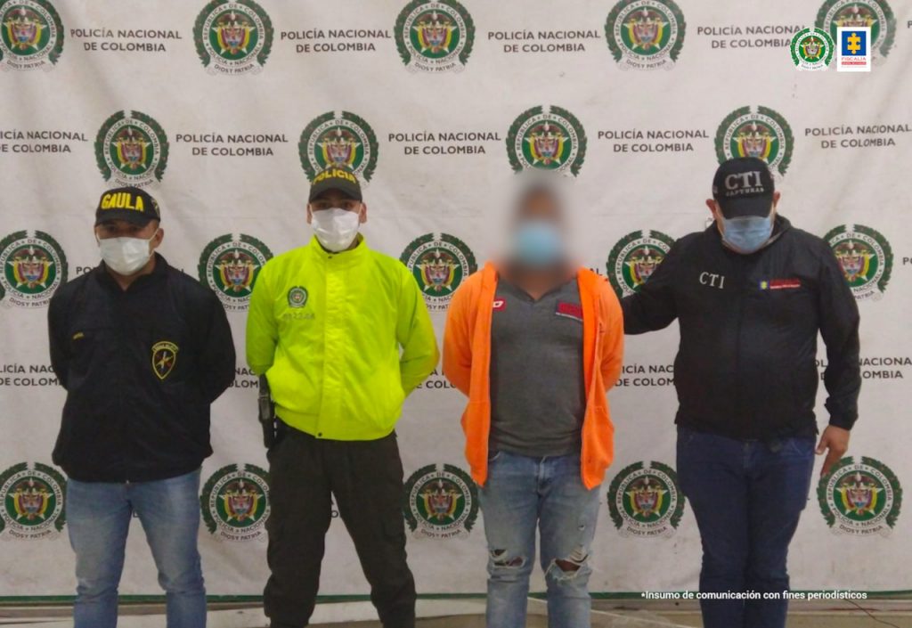 En la fotografía aparece un hombre de camiseta gris con chaqueta naranja, custodiado por uniformados del CTY la Policía Nacional, delante de un pendón de la Policía Nacional.