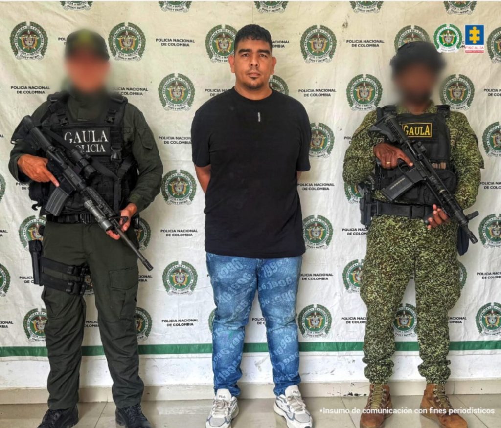 En la fotografía está un hombre trigueño de pie, con los brazos atrás esposados, trigueño, cabellos negros lisos y cejas gruesas negras, tiene una semi barba y está vestido con camiseta negra, jean azul y tenis blanco. Se encuentra acompañado de un uniformado de la Policía y otro del Ejército Nacional.