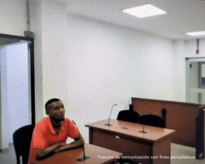En la imagen está un hombre delgado, moreno, cabellos negros, vestido con camiseta roja, se encuentra sentado en un escritorio de madera con un micrófono, en la sala de audiencia donde se le procesó.