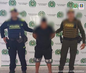 En la imagen está un hombre de pie con el rostro difuso, vestido de camiseta y bermuda negra, custodiado por dos agentes de la Policía Nacional. Detrás de ellos hay un pendón de esa institución.