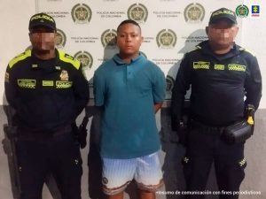  En la imagen está un hombre moreno, de pie, con los brazos atrás, de contextura gruesa, vestido de camiseta azul y bermuda azul con café, acompañado de dos uniformados de la Policía Nacional. 