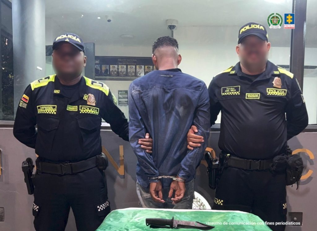 En la fotografía hay un hombre de pie, alto, moreno, está de espalda, cabellos cortos negros crespos, tiene los brazos esposados y está vestido con una camisa manga larga azul. Se encuentra acompañado por dos policías.