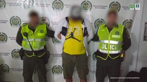 En la imagen se encuentra un hombre de pie con los brazos atrás y el rostro difuso vestido con una camiseta amarilla, saco blanco y bermuda verde. Se encuentra acompañado por dos uniformados de la Policía Nacional.