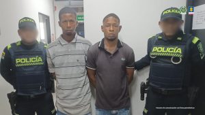 En la foto están dos hombres de pie, con los brazos atrás, jóvenes, delgados, morenos, con cabellos cortos negros, uno se encuentra vestido con una camiseta gris y rallas negras y el otro con una camiseta gris oscura. Están acompañados por dos agentes de la Policía Nacional.