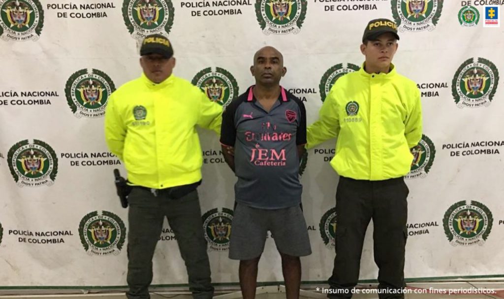 En la fotografía se observa a un hombre de camiseta y bermuda oscura, custodiado por dos agentes de la Policía Nacional, delante de un pendón de esa institución.