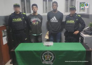 En la imagen aparecen dos hombres de pie, con los brazos atrás, vestido de buzos negros, custodiados por dos uniformados de la Policía Nacional. Detrás de ellos hay un pendón de esa institución y delante de ellos una mesa con un reloj.