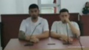 En la fotografía están dos hombres sentados frente a un escritorio de madera, morenos, ambos vestidos con camisetas blancas, tienen micrófonos y se encuentran al interior de la salda de audiencia donde se le procesó. 