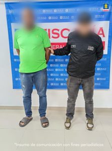 En la imagen está un hombre de pie, de frente, alto, contextura mediana, con los brazos atrás esposados, tiene el rostro difuminado y se encuentra vestido con camiseta verde, jean azul y sandalias grises. A su lado está un uniformado del CTI y detrás de ellos hay una bandera de la Fiscalía.