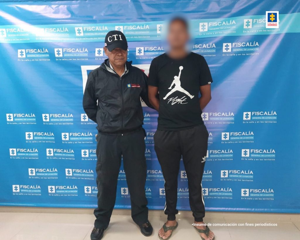 En la imagen aparece un hombre de tez morena, vestido con camiseta, sudadera negra y chancletas negras, con los brazos atrás, custodiado por un uniformado del CTI. Detrás de ellos hay un pendón azul que dice Fiscalía General de la Nación.