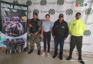 En la fotografía está la imagen de una mujer joven, blanca, delgada, vestida de camiseta gris con jean negro y sandalias blancas. A su lado se encuentran uniformados del CTI, Ejército y Policía Nacional.