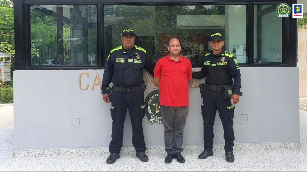 En la imagen aparece un hombre de tez blanca, semi calvo, vestido de camiseta roja con jean azul, custodiado por dos agentes de la Policía Nacional en una estación de esa institución.
