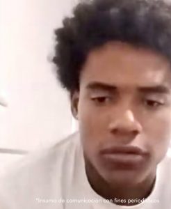  En la fotografía está el rostro de un hombre joven, moreno, de cabellos largos crespos, nariz y boca grande, se encuentra vestido con una camiseta blanca y está sentado en el interior de una oficina asistiendo virtualmente a la audiencia en la que se le procesó. 