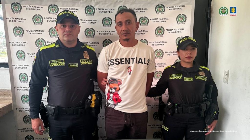 En la fotografía aparece un hombre de pie, con los brazos atrás esposados, trigueño, con la frente despoblada, vestido con camiseta blanca y pantalón café. Se encuentra custodiado por dos uniformados de la Policía Nacional.