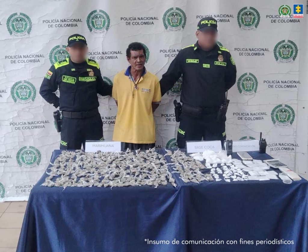 En la fotografía está un hombre de pie, de frente, moreno, delgado, de cabellos negros, de avanzada edad, vestido con una camiseta amarilla y se encuentra custodiado por dos uniformados de la Policía Nacional. Delante suyo hay una mesa con las bolsitas de drogas incautadas. 