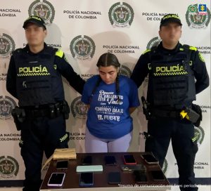 En la fotografía está una mujer trigueña, de contextura mediana, de baja estatura, cabellos largos negros, trenzados con la cabeza agachada y los brazos atrás esposados. Está vestida con una camiseta azul oscura y un pantalón azul claro. Delante de ellas hay una mesa con celulares incautados.