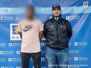  En la fotografía hay un hombre con el rostro difuso vestido de camiseta rosada con jean negro custodiado por un funcionario del CTI. Detrás de ellos hay un pendón que dice Fiscalía General de la Nación.