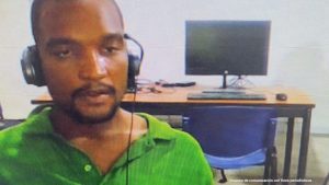 En la imagen está un hombre moreno con barba, labios gruesos, semi calvo, vestido con camiseta verde. Tiene unos audífonos y se encuentra al interior de una oficina asistiendo virtualmente a la audiencia en la que se le procesó.