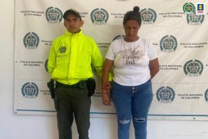 En la fotografía se observa a una mujer vestida de jean con camiseta blanca, custodiada por un agente de la Policía Nacional. Detrás de ellos se divisa un pendón de esa institución.