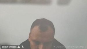 En la imagen está el medio rostro de un hombre trigueño, semi calvo, cabellos negros, se encuentra con la cabeza agachada, vestido con una chaqueta negra y está sentando en el interior de una oficina asistiendo virtualmente a la audiencia en la que se le procesó. 