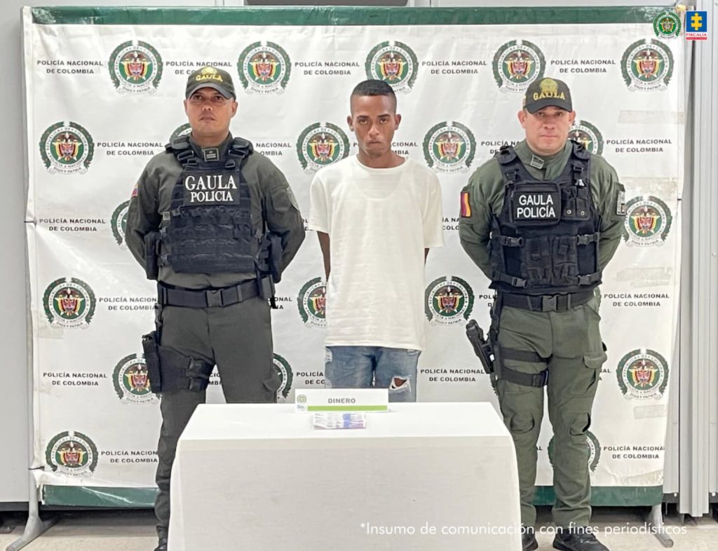 En la imagen está un hombre delgado de pie, de frente, moreno, con cabellos cortos, los brazos atrás esposados, vestido con una camiseta blanca y jean azul, custodiado por dos uniformados de la Policía Nacional. Delante de ellos hay una mesa con el dinero incautado.