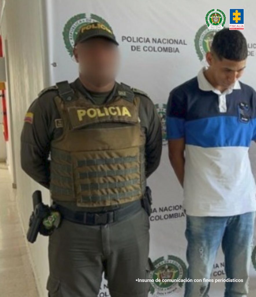 En la fotografía está un hombre de pie, con los brazos atrás, delgado, vestido de camiseta azul con rallas blancas y jean azul, acompañado de un uniformado de la Policía Nacional.