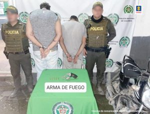 En la imagen aparecen dos hombres de espalda con las manos esposadas, ambos vestidos de camisetas y pantalón gris, custodiado por dos agentes de la Policía Nacional. Delante de ellos hay una mesa donde se encuentra el arma que se le incautó. Detrás de ellos está un pendón de la Policía Nacional.