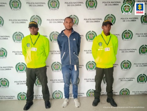 En la fotografía se observa a un hombre de tez morena vestido de buzo azul con jean y tenis blanco, custodiado por dos agentes de la Policía Nacional, delante de un pendón de esa institución.