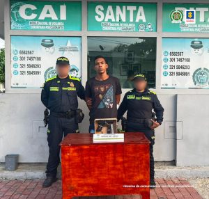 En la imagen hay un hombre de pie con brazos atrás, vestido de camiseta café con jean azul. Se encuentra acompañado de dos uniformados de la Policía Nacional. Delante de él en una mesa está el arma que se le incautó.
